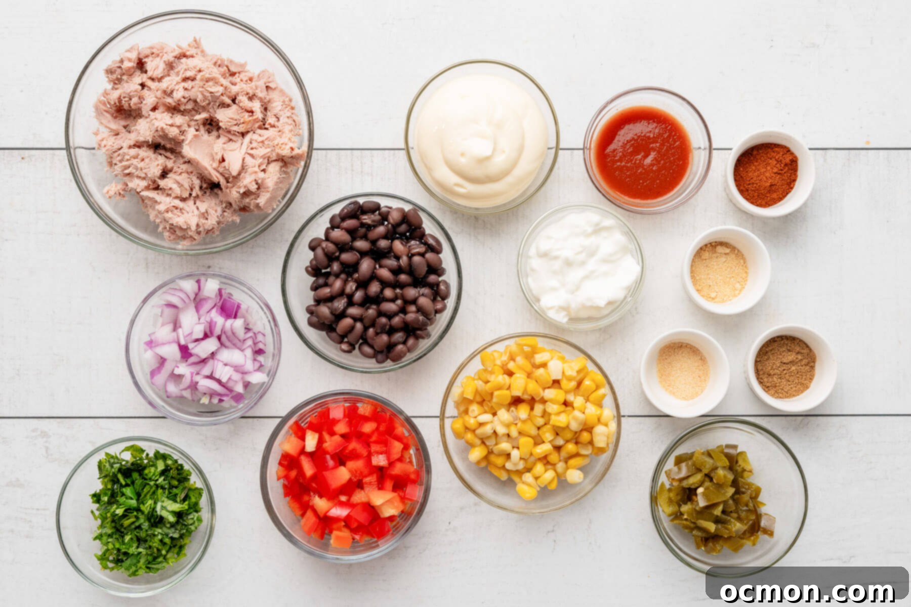 All the fresh ingredients for Mexican Tuna Salad, including drained tuna, corn, black beans, bell pepper, red onion, cilantro, and pickled jalapeños, neatly separated in small bowls on a wooden surface.