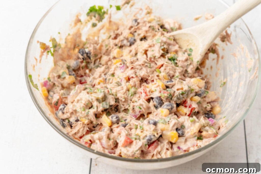 The Mexican Tuna Salad ingredients and dressing are thoroughly mixed together in a large bowl with a wooden spoon, creating a creamy and colorful mixture.