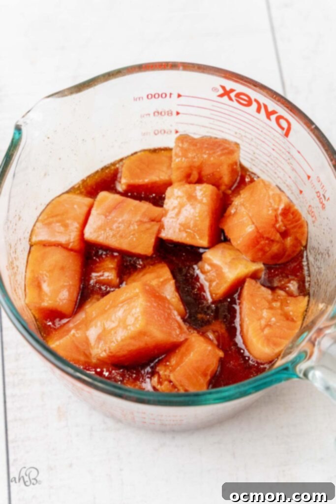 Uniformly cubed salmon pieces are submerged and marinating in a rich, savory hot honey and soy sauce mixture inside a clear glass measuring bowl, infusing them with flavor.