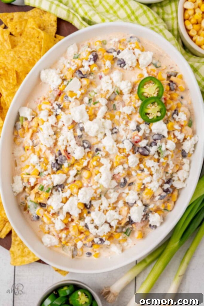 A white serving bowl brimming with colorful Fiesta corn dip, generously topped with crumbled queso fresca cheese and slices of fresh green jalapeno pepper. 