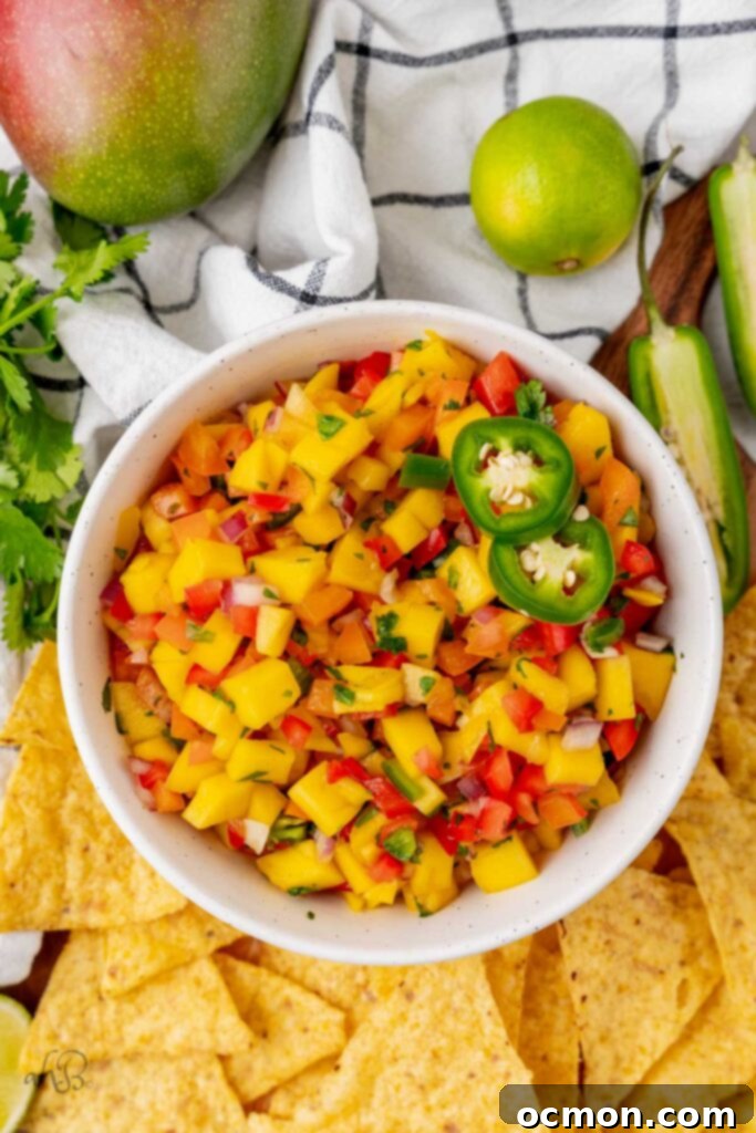 A white bowl of spicy mango salsa with a pile of tortilla chips to one side and limes to the other. 