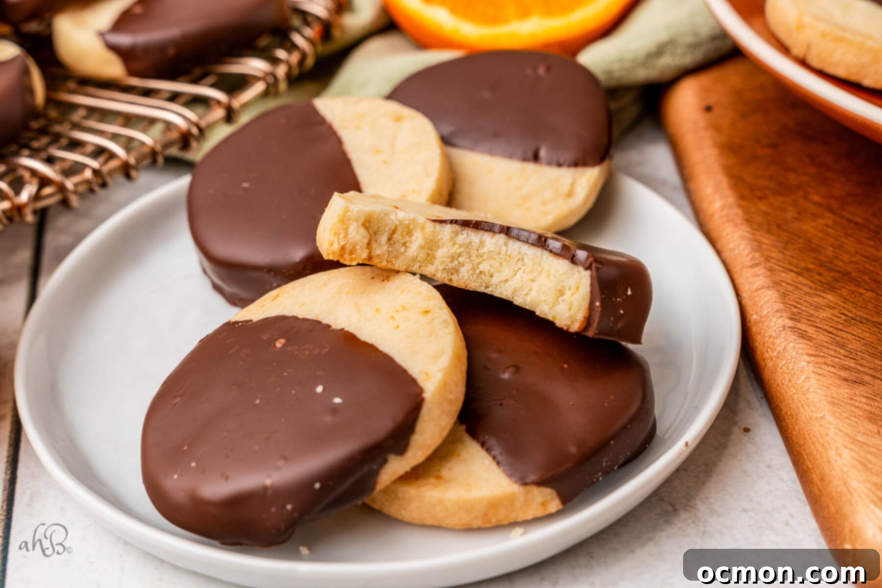 Orange Zest Chocolate Shortbread 2 A white plate of chocolate Orange Shortbread Cookies with a bite taken out of the top cookie, showcasing the perfect orange-infused shortbread and dark chocolate coating.