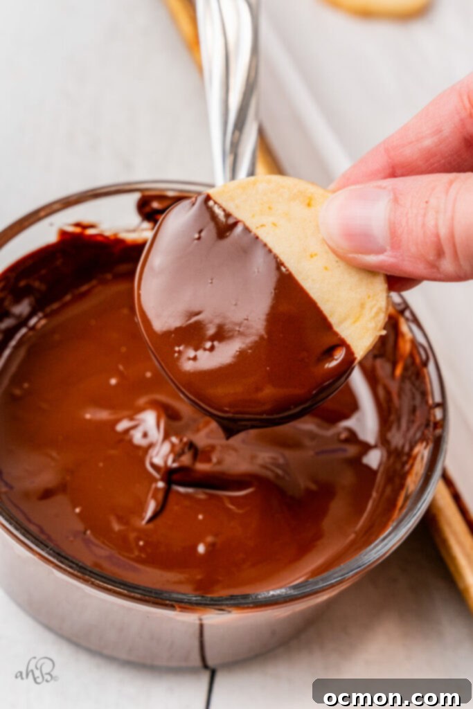 Orange Zest Chocolate Shortbread 12 A delicate shortbread cookie is being artfully dipped halfway into a glass bowl filled with rich, melted dark chocolate, creating a striking contrast.