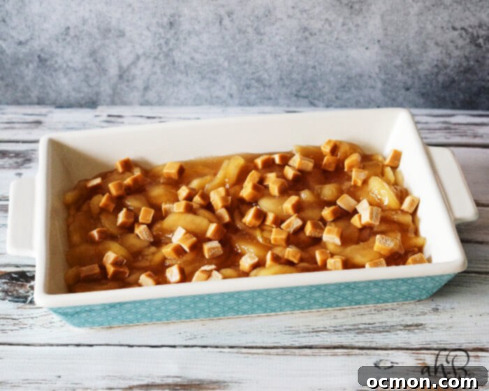 Small, chopped caramel pieces are generously sprinkled over the vibrant apple pie filling layer in the baking dish.
