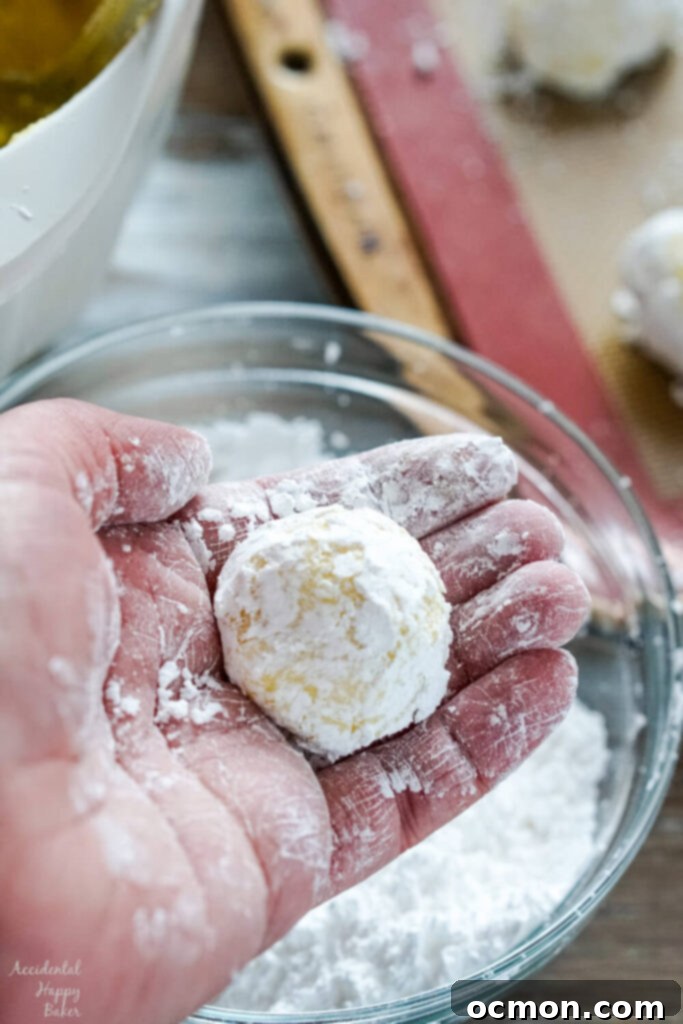 Step two: rolling the sticky lemon cookie dough into small, uniform balls, and then coating each ball generously in powdered sugar for the characteristic crinkle effect.