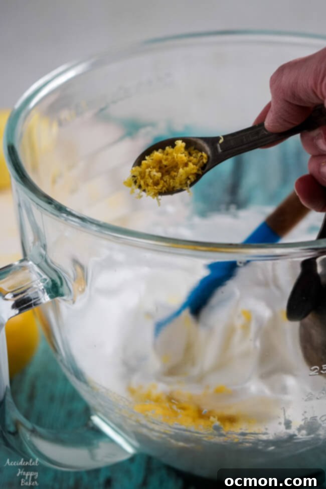 A hand adding finely grated fresh lemon zest to the fluffy white meringue batter in a mixing bowl.