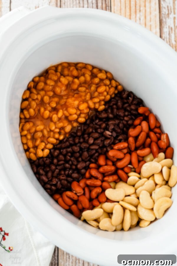 Canned beans, including butter beans, kidney beans, and black beans, along with ground beef and bacon, in a slow cooker, ready for the sauce to be added.