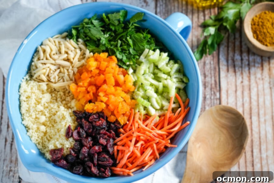 Golden Curried Couscous Salad 3 A blue bowl filled with separate piles of raw ingredients for curried couscous salad, including golden couscous, sliced almonds, fresh parsley, chopped celery, shredded carrots, craisins, and dried apricots, ready for mixing.