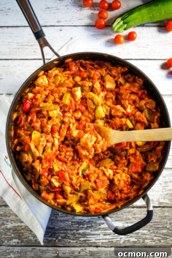 A skillet full of Italian Chicken and Orzo with a wooden spoon and zucchini and tomatoes in the background. Close-up of a rustic, hearty one-pot chicken and orzo dish, perfect for a quick dinner.