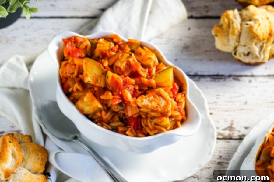 A white bowl full of Italian Chicken and Orzo sits next to a few garlic rolls. This hearty one-pot meal is perfect for busy weeknights, offering a complete and satisfying dinner.