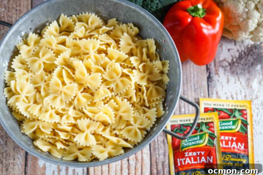 Vibrant Summer Veggie Pasta 4 A colander of perfectly cooked farfalle pasta sits next to a vibrant pile of chopped vegetables including bell pepper, cucumber, broccoli, and red onion, with two packets of Italian dressing mix nearby.