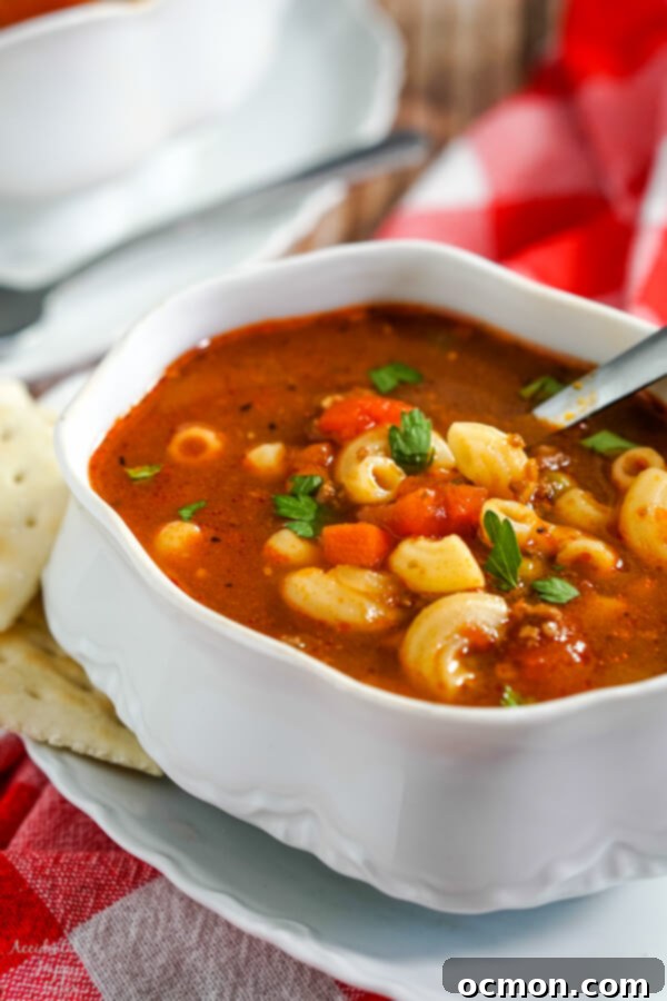 A white soup tureen full of hamburger macaroni soup with a soup spoon in the bowl and crackers to the side.