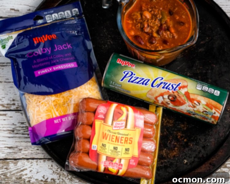 The Chili Dog Pie 3 All the essential ingredients for Chili Dog Pizza laid out: a prepared pizza crust on a pan, a bowl of shredded cheese, a bowl of sliced hot dogs, and a container of savory chili, ready for assembly.