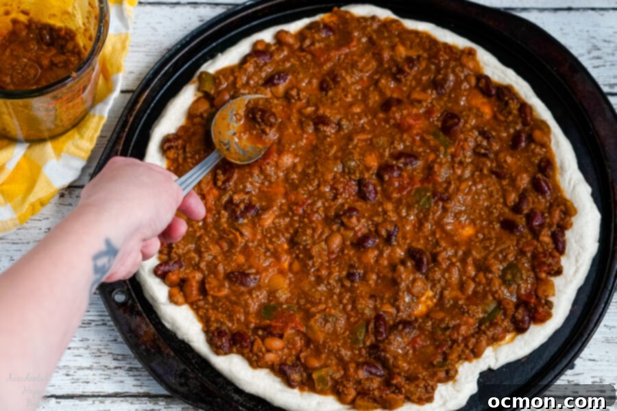 The Chili Dog Pie 4 Thick, hearty chili is spread evenly across the surface of a pre-baked pizza dough, acting as the flavorful base sauce for the Chili Dog Pizza.