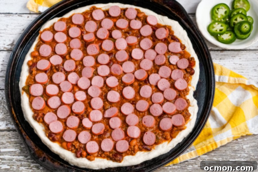 The Chili Dog Pie 5 Neatly sliced hot dog pieces are arranged over the chili-covered pizza dough, creating a visually appealing and delicious layer before the cheese is added.