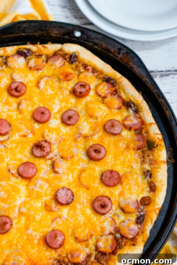 The Chili Dog Pie 7 A close-up of a chili dog pizza slice, showing the melted cheese, chili, and hot dog slices, garnished with fresh chopped onions, highlighting its readiness to be enjoyed.