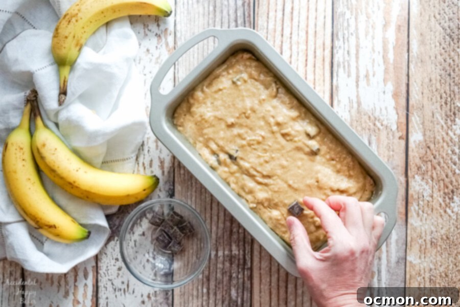 A loaf pan full of banana bread batter as more chocolate chunks are added to the top. 