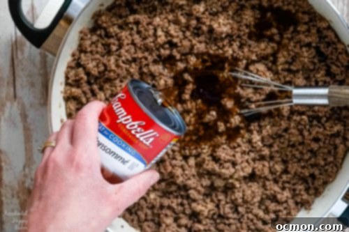 Ground beef mixture simmering in a skillet with beef consommé and Worcestershire sauce, creating a rich, savory sauce.