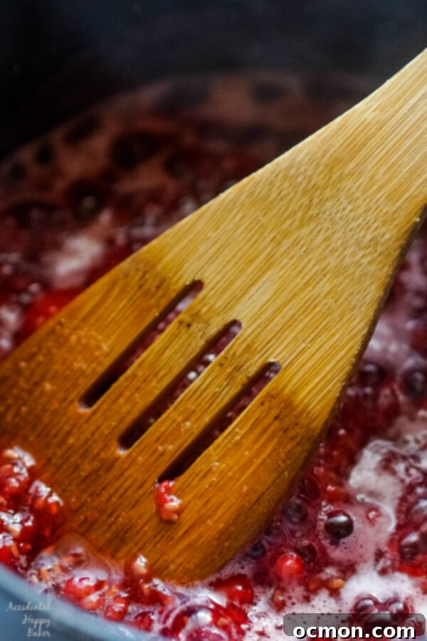 Whipped Raspberry Honey Butter 5 A close-up shot of vibrant red raspberries being cooked and vigorously mashed with a wooden spoon in a saucepan with honey, creating a flavorful syrup.