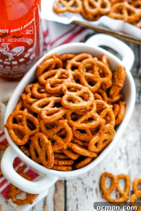 A large white bowl filled with golden-brown spicy sriracha ranch pretzels, with a bottle of sriracha sauce resting beside it, highlighting the key ingredient.