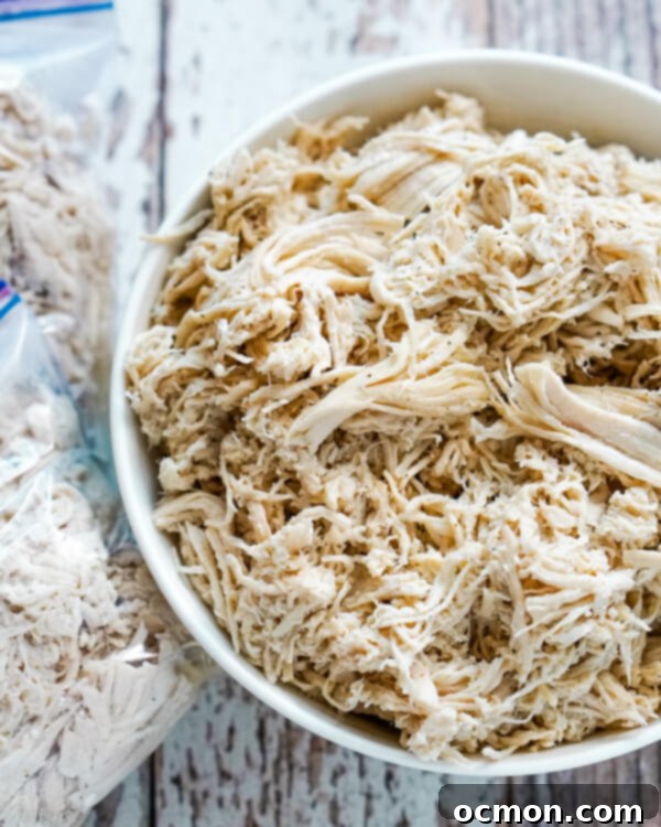 A white bowl full of shredded chicken with 2 freezer bags of chicken next to it.