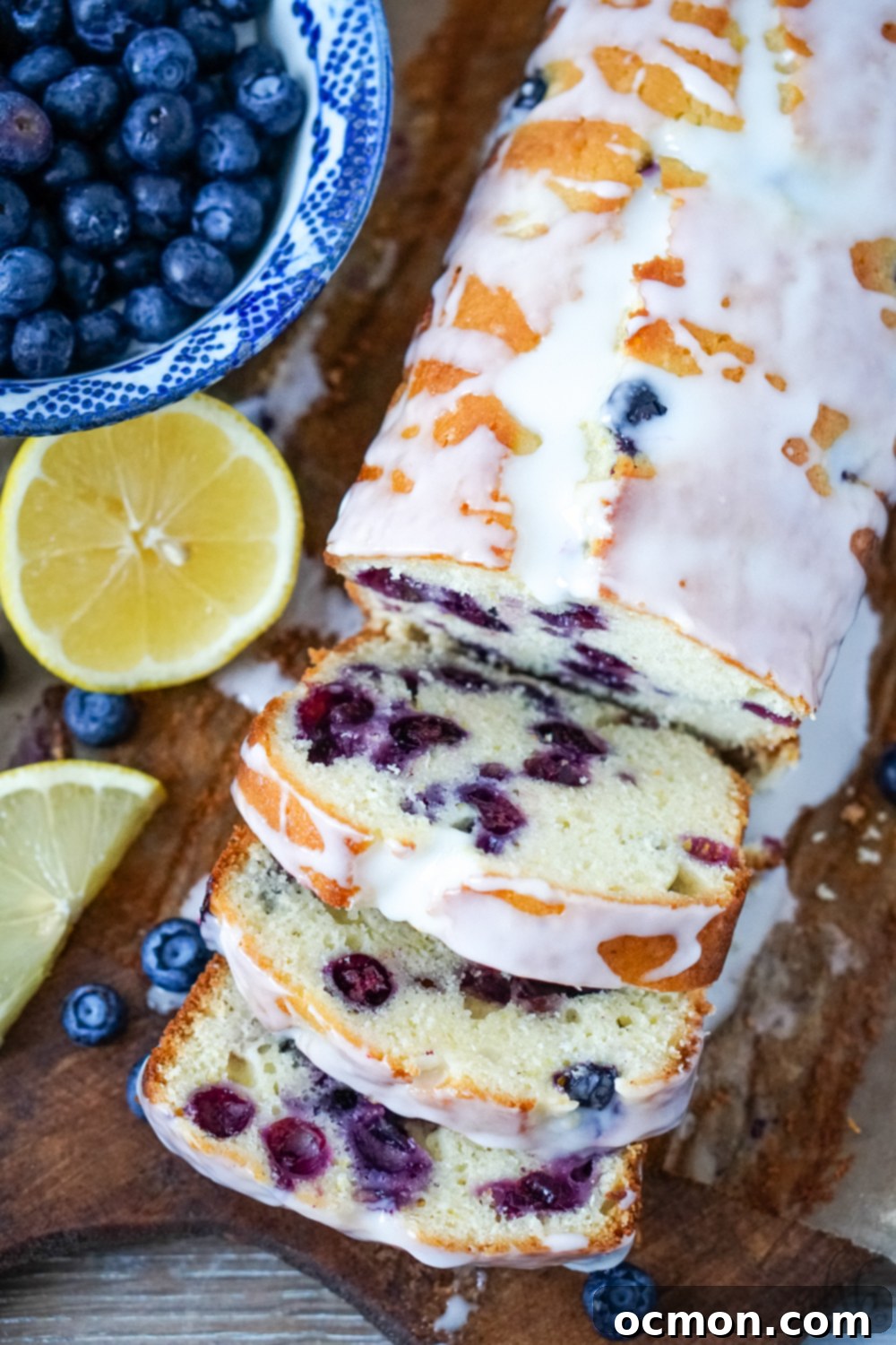 Zesty Lemon Blueberry Loaf with Creamy Buttermilk Glaze 2 A beautifully sliced loaf of lemon blueberry bread with buttermilk glaze drizzled over the top, resting on a rustic bread board alongside fresh lemon slices and a bowl of vibrant blueberries.