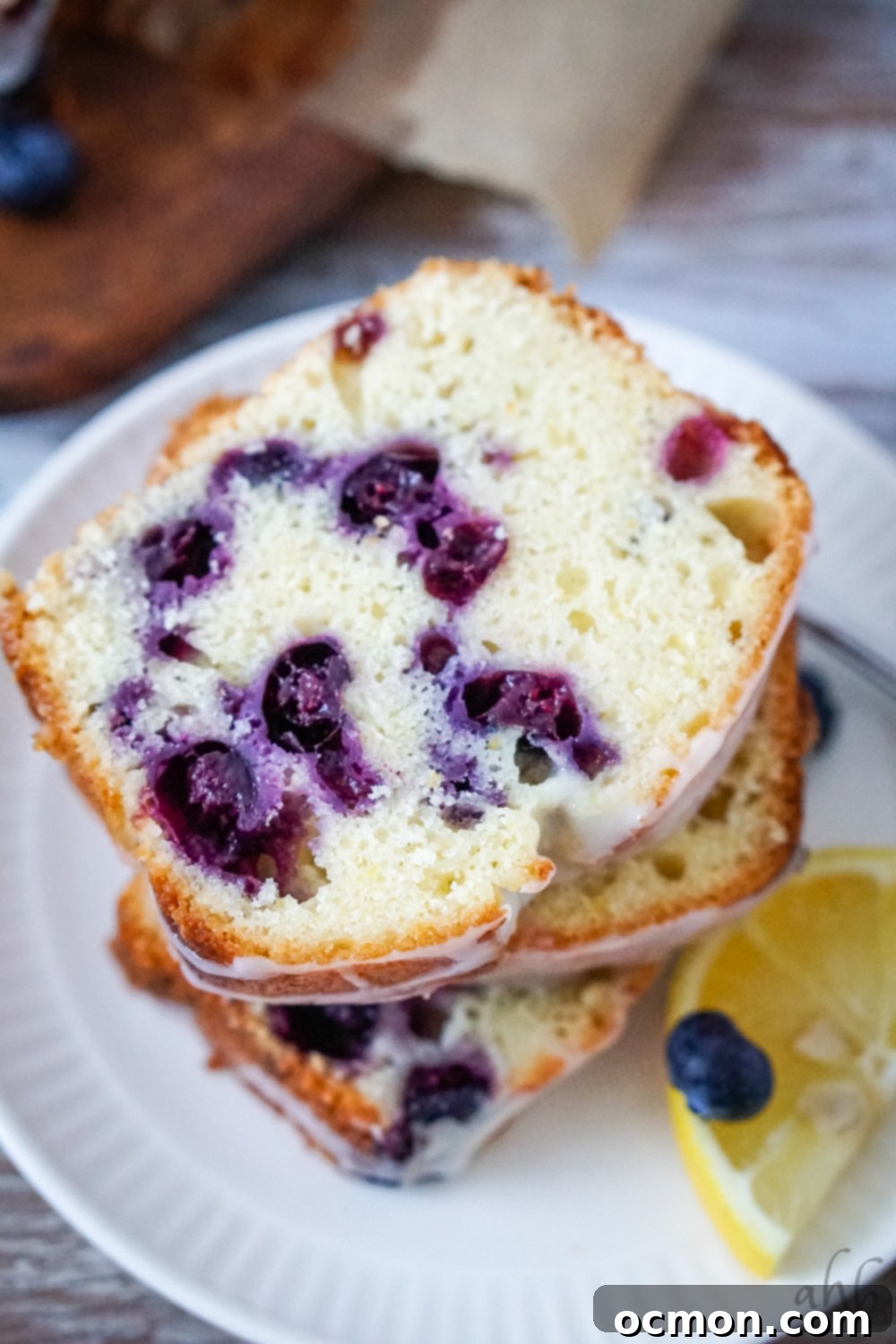 Zesty Lemon Blueberry Loaf with Creamy Buttermilk Glaze 3 Several moist slices of lemon blueberry bread, speckled with blueberries, arranged elegantly on a white plate with a fresh slice of lemon for garnish.