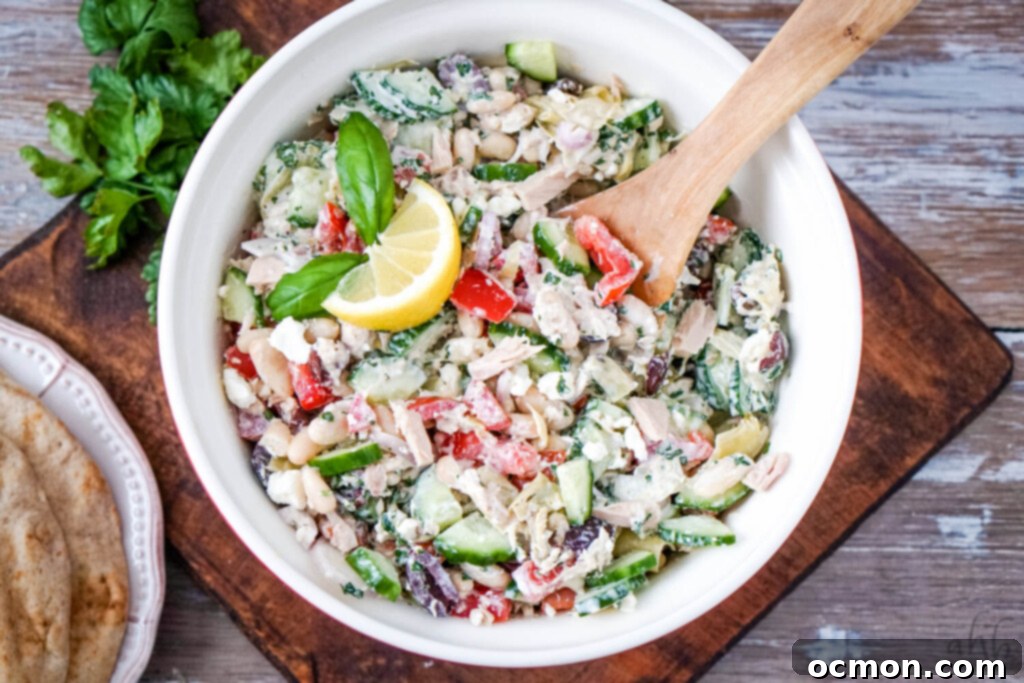 A vibrant red bowl filled with Mediterranean Tuna Salad, garnished with fresh herbs, with a stack of warm pita breads on the side.
