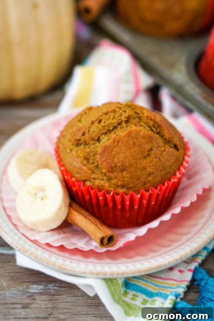 A perfectly baked Pumpkin Banana Muffin, still warm, rests in its paper liner on a decorative white plate, accompanied by two fresh banana slices and a fragrant cinnamon stick, highlighting its rich, spiced appeal.