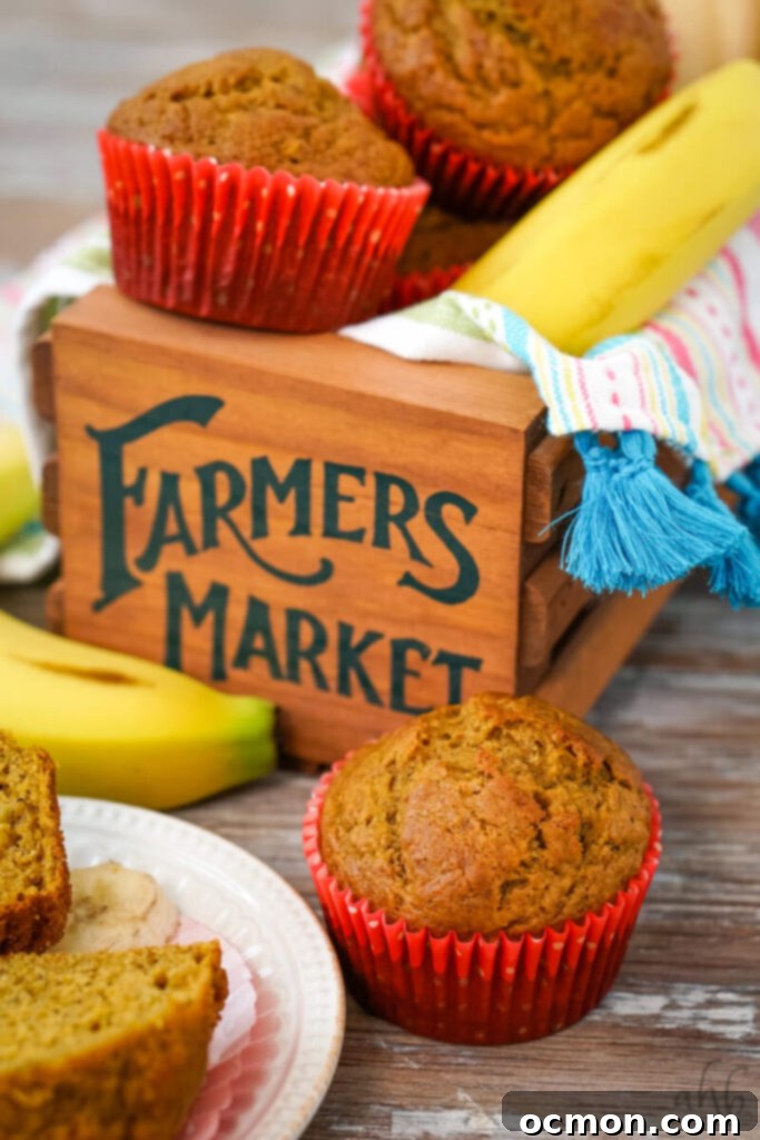 A close-up shot of a plump Pumpkin Banana Muffin in a vibrant red paper liner, alongside a sliced banana. In the background, two more muffins in red liners and a banana rest on a rustic wooden box labeled 'Farmers Market', suggesting fresh, wholesome ingredients.