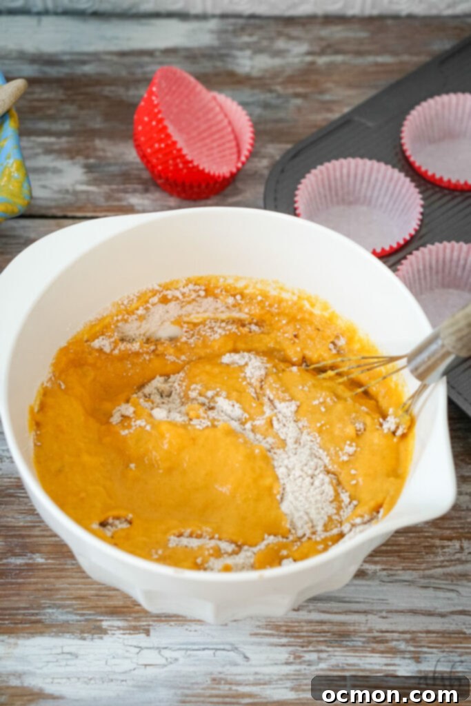 The smooth, vibrant orange-hued wet mixture is carefully poured into a well created in the center of the dry ingredients in the white mixing bowl, then gently stirred with a whisk until just combined, showing a slightly lumpy but moistened batter.