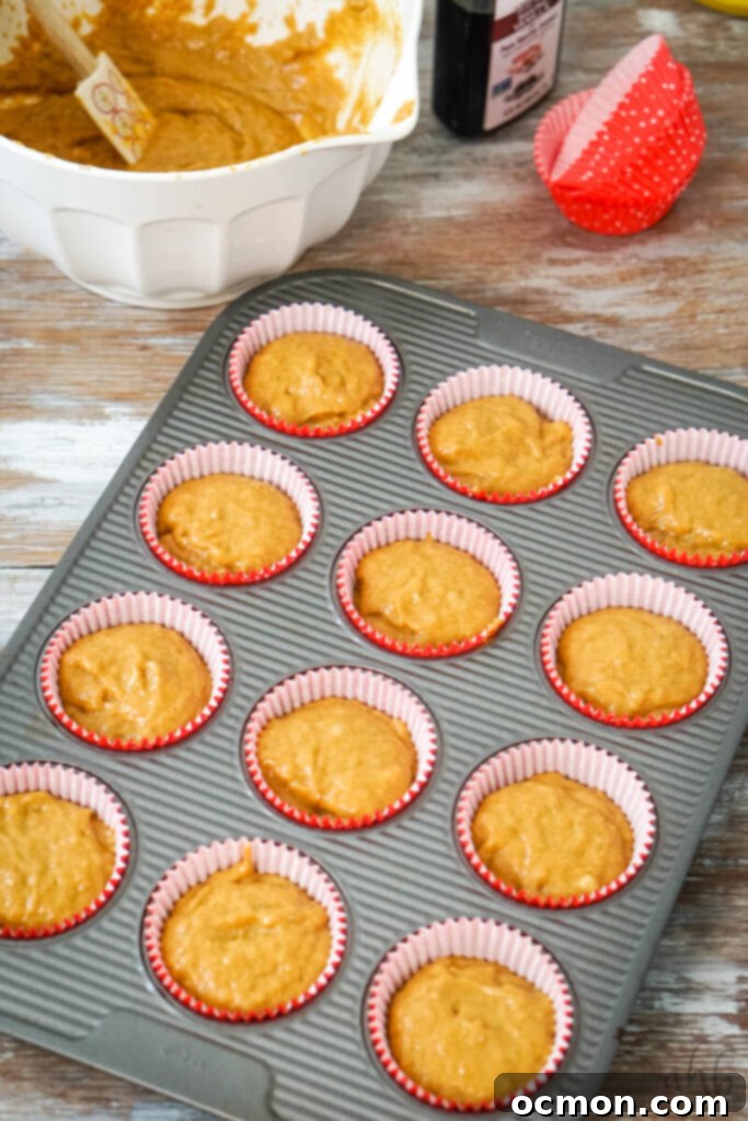 The thick, spiced pumpkin banana muffin batter is neatly scooped and evenly distributed into twelve muffin cups, each lined with a festive paper muffin liner, filling them approximately three-quarters full.
