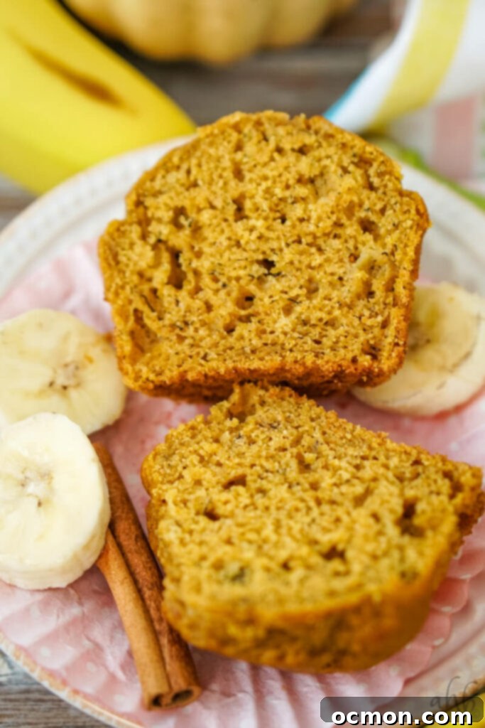 A freshly baked, domed pumpkin banana muffin, unwrapped and halved, showcases its perfectly moist and spiced interior, positioned beside fresh banana slices and aromatic cinnamon sticks, highlighting its delicious components.