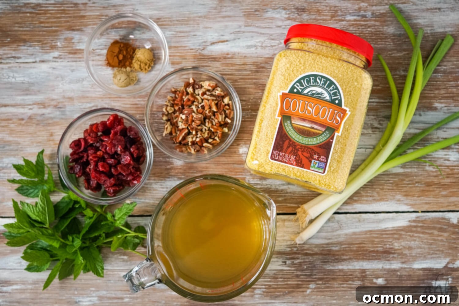 A curated display of key ingredients for Cranberry Couscous Salad: clear bowls holding cinnamon, cumin, and cardamom, pecan pieces, and dried cranberries; fresh mint leaves, a glass measuring cup with homemade dressing, a container of Rice Select Couscous, and three green onions arranged on a rustic wooden table, ready for preparation.