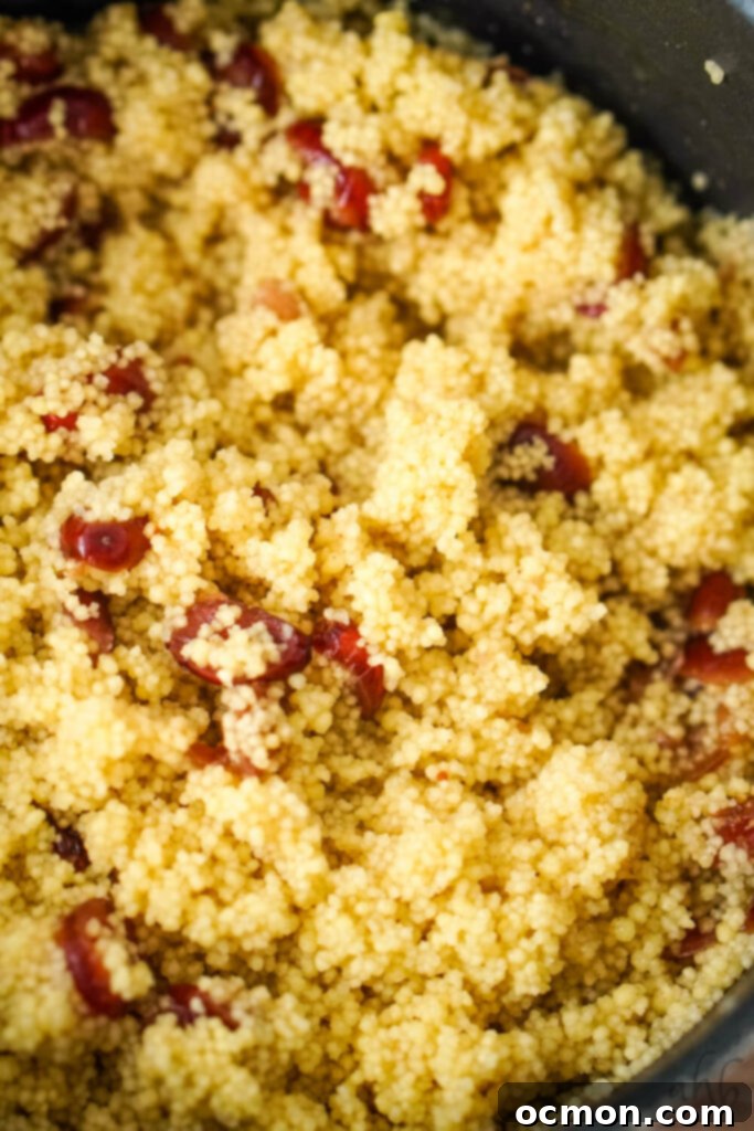 Cooked couscous mixed with the dried cranberries, cinnamon, cardamom, and cumin, steeping in a saucepan with the lid slightly ajar after boiling.