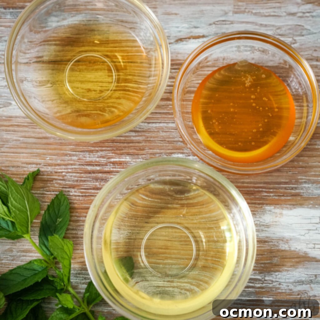 Three clear glass bowls containing the separate dressing ingredients: vegetable oil, honey, and apple cider vinegar, positioned on a wooden surface.