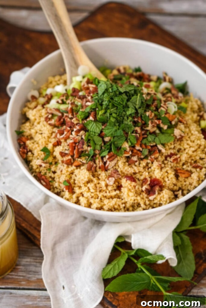 A white bowl of chilled cranberry couscous, with toasted pecans, chopped green onions, and fresh mint leaves being gently stirred with a wooden spoon, ready for the dressing.