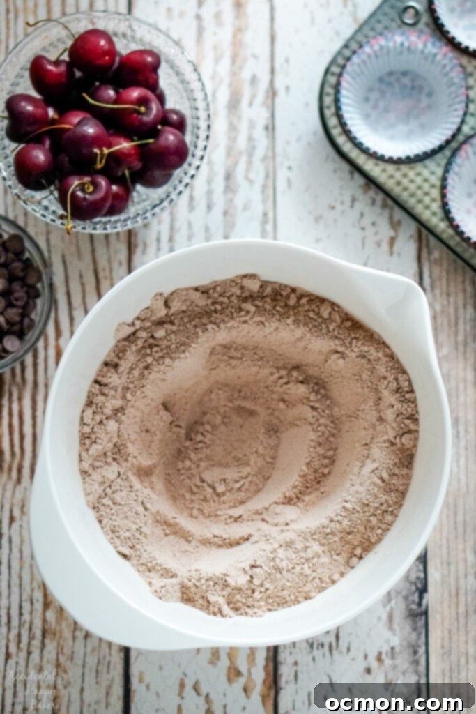 A vibrant collection of baking ingredients: a bowl of pitted cherries, a bowl of dark chocolate chips, a muffin tin lined with paper cups, and a large bowl holding the perfectly mixed dry muffin ingredients, ready for the next step.