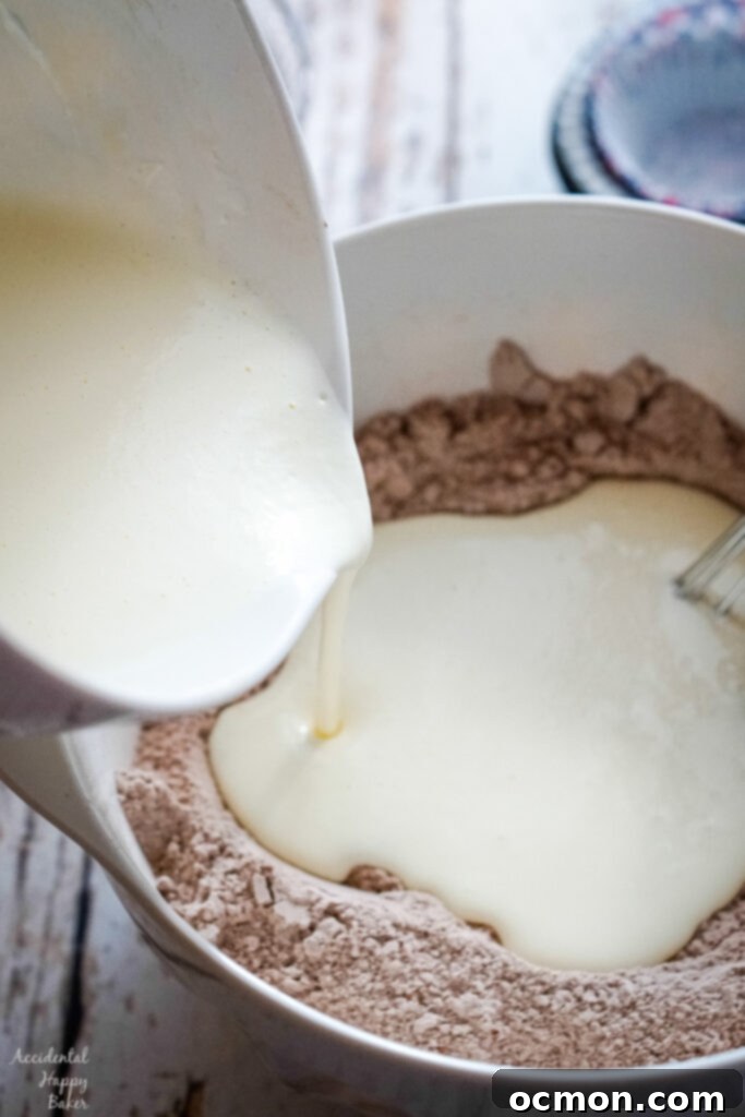 The process of mixing: a smooth, liquid mixture of wet ingredients (sour cream, buttermilk, oil, eggs, almond extract) being carefully poured into a central well formed in the dry ingredients, ready to be gently whisked together.