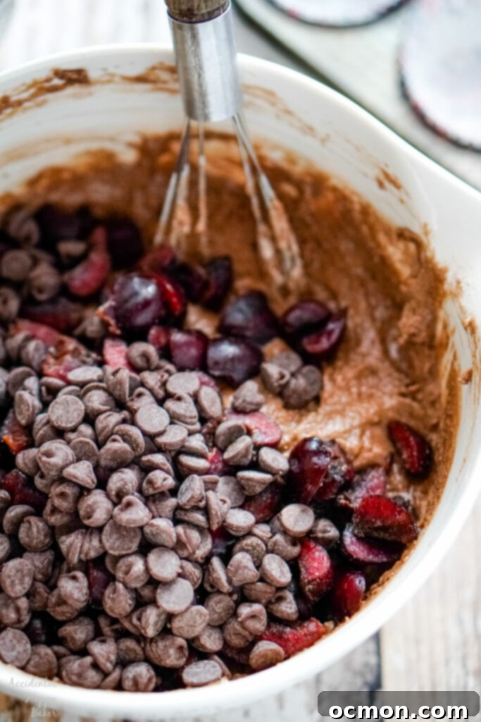 A close-up shot of chopped, juicy cherries and glistening dark chocolate chips being carefully folded into the rich, chocolatey muffin batter, ensuring even distribution of these key ingredients.