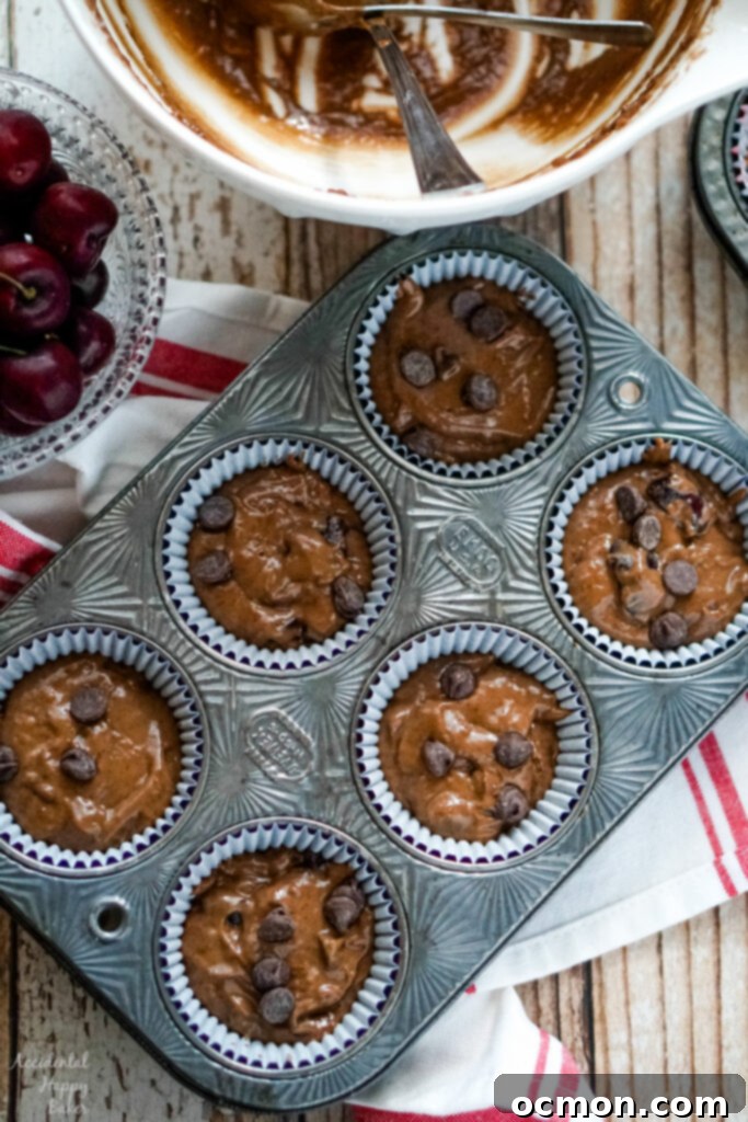 Rich chocolate cherry muffin batter being carefully scooped into paper-lined muffin cups, filling them approximately two-thirds full, ready for baking.
